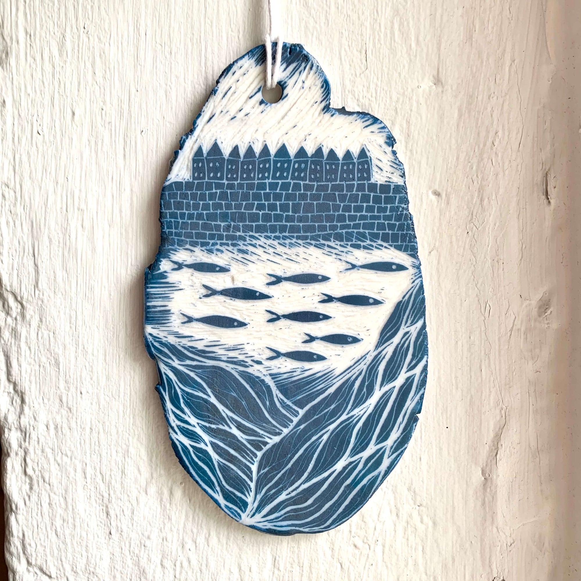 Handcrafted by Sarah Livingstone. White and blue porcelain rough oval shaped hanging, with waves, fishes and seaside town carved into it.