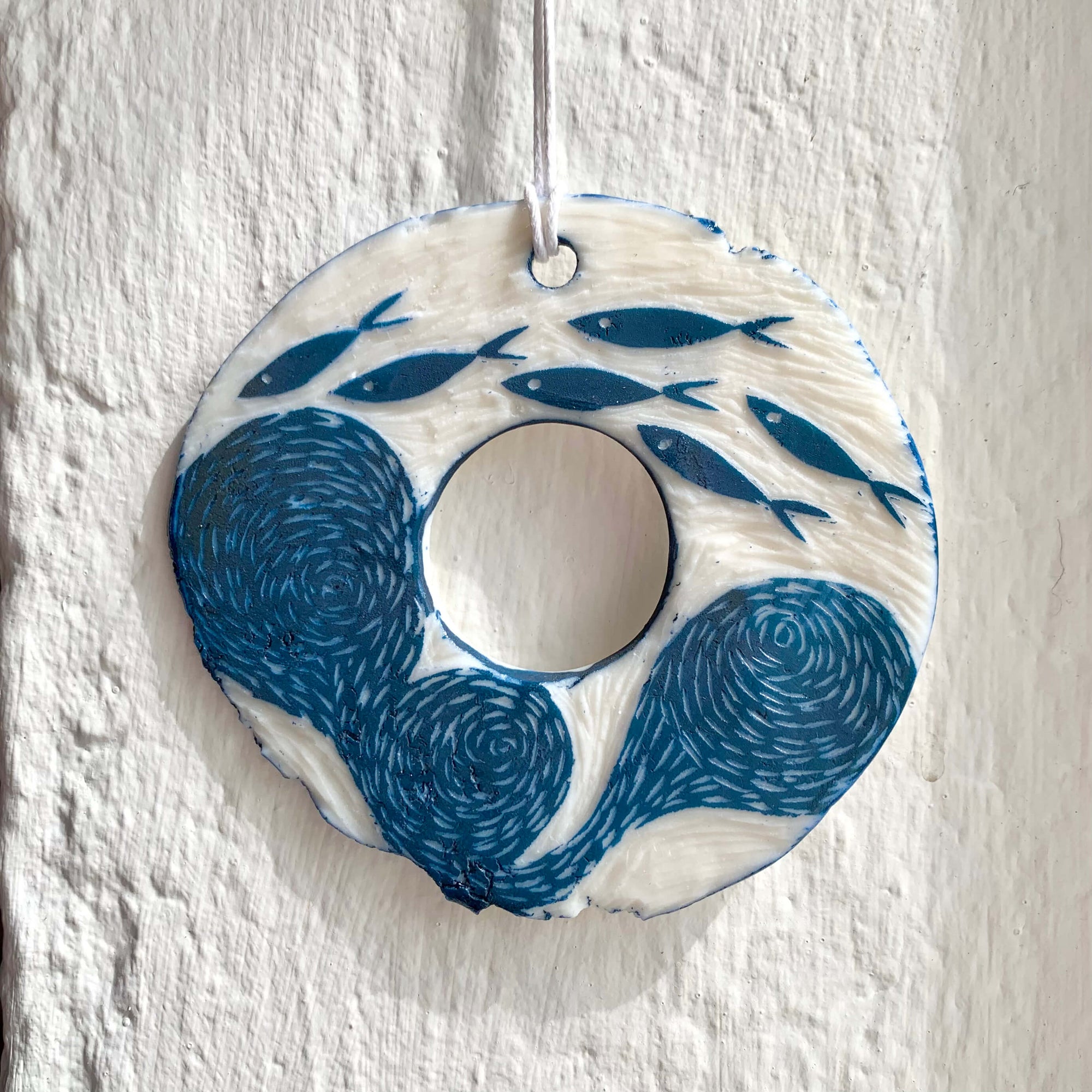 Handcrafted by Sarah Livingstone. White and blue porcelain rough circular shaped hanging, with a hole in the middle and swirly waves and fishes carved into it.
