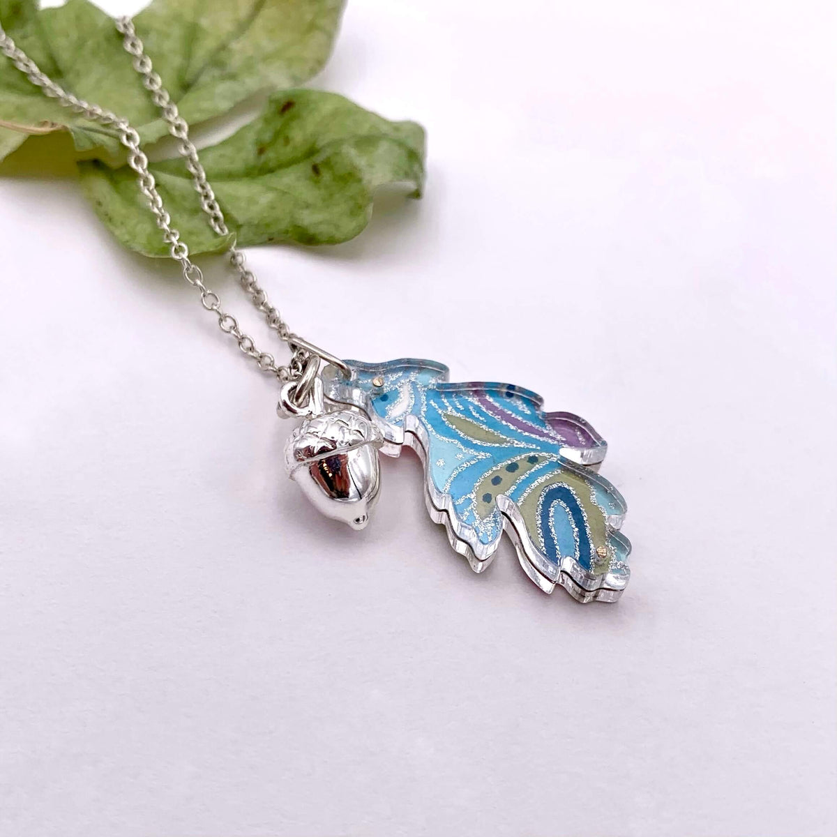 Handmade silver chain necklace, featuring a solid silver acorn and colourful reversible leaf. Leaf is made from blue floral hand-painted paper, between two layers of perspex.