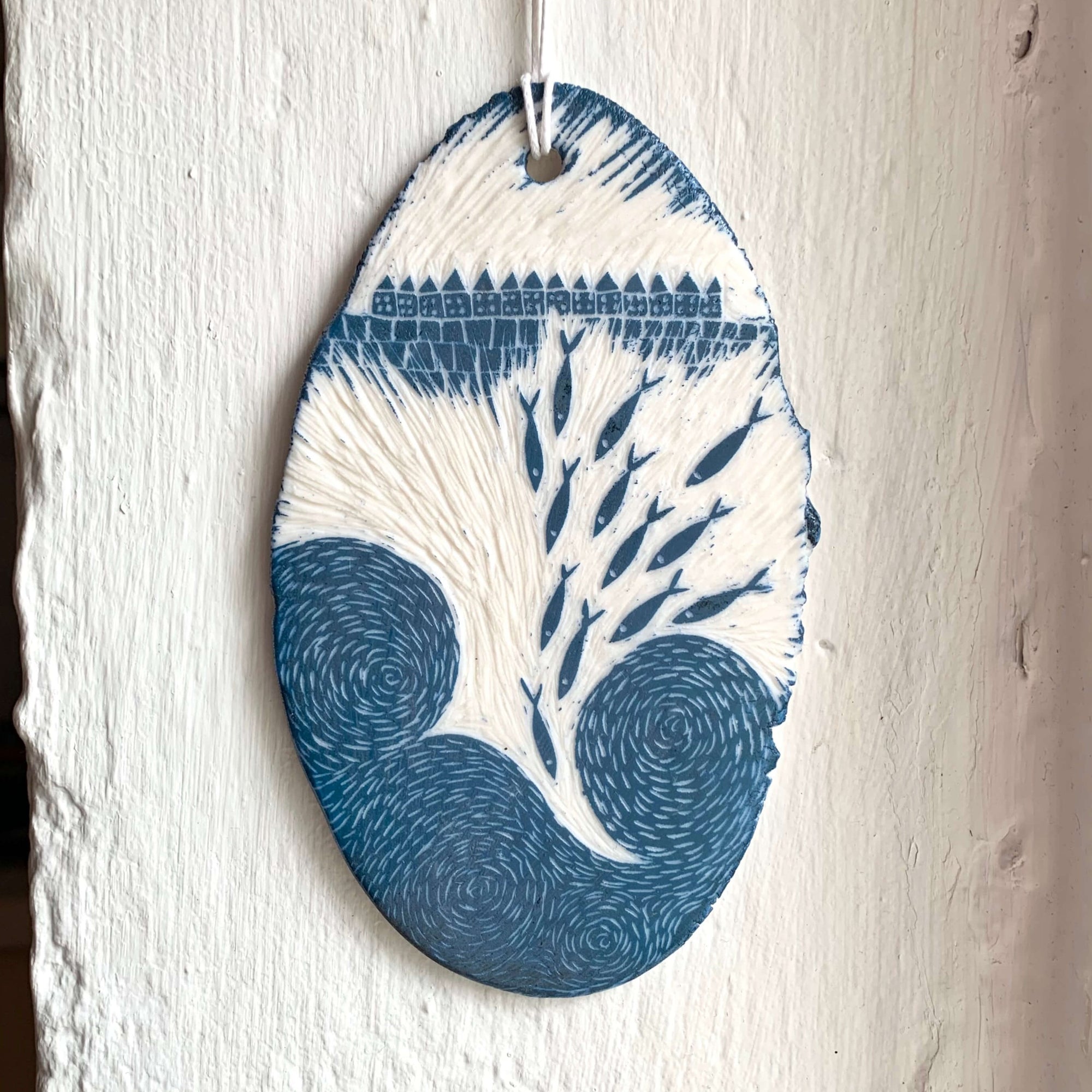 Handcrafted by Sarah Livingstone. White and blue porcelain rough oval shaped hanging, with abstract waves, fishes and small buildings carved into it.