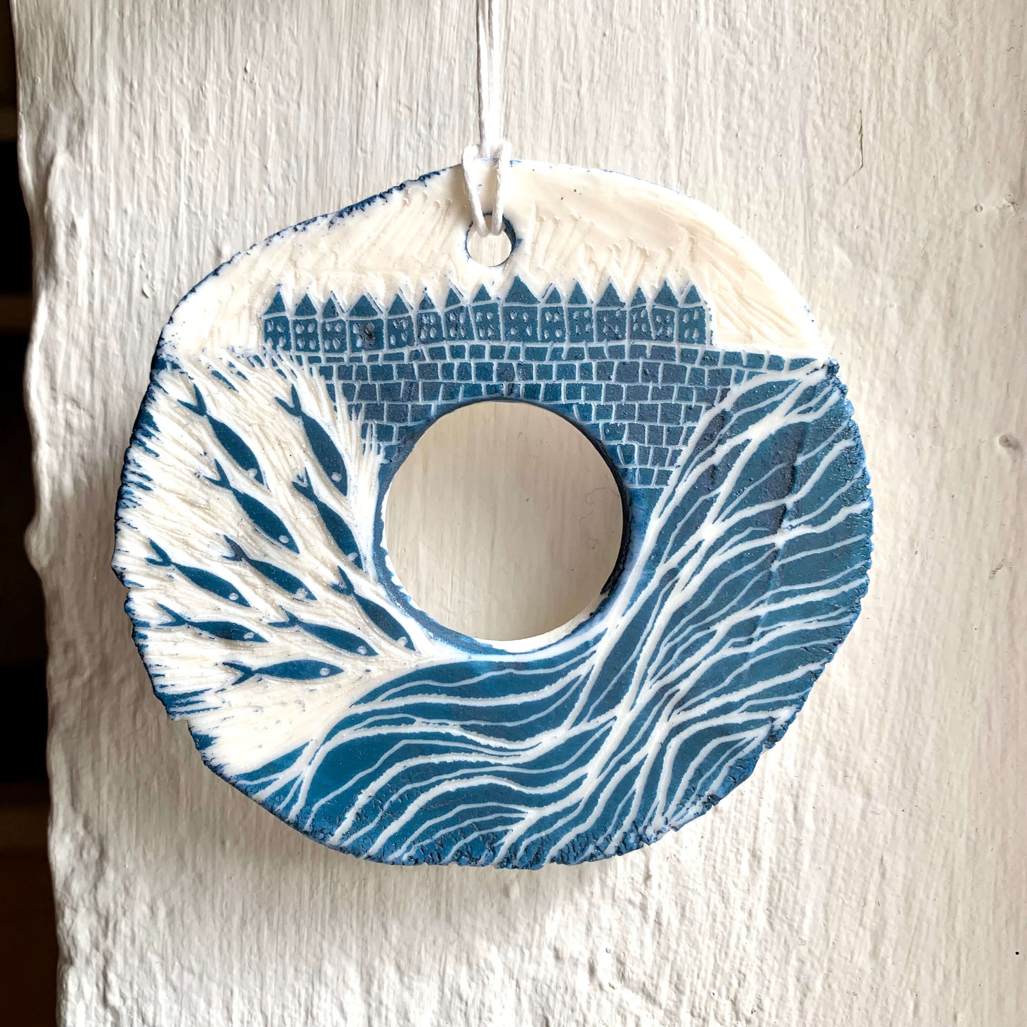 Handcrafted by Sarah Livingstone. White and blue porcelain rough circular shaped hanging, with a hole in the middle and abstract waves, fishes and small buildings carved into it.