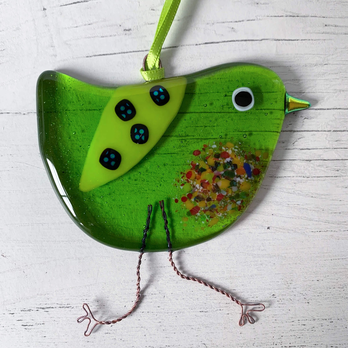 Handmade Fused Glass Green Hanging Bird, on a silky green ribbon. Featuring a multicoloured textured belly, wire legs, an eye and patterned wing. By Sarah Myatt in Staffordshire.
