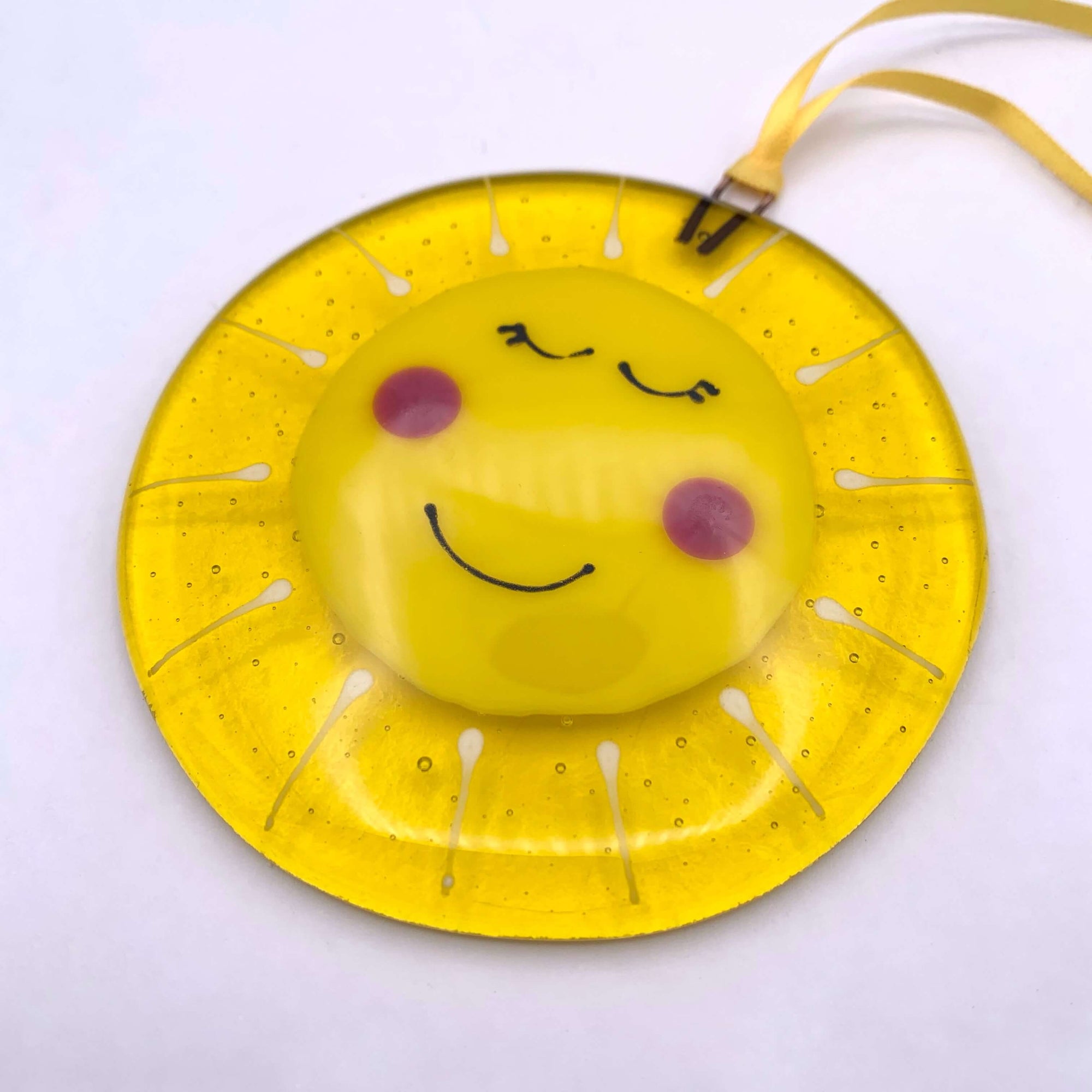 Handmade fused glass yellow circle hanging, with a smiley faced sunshine on the front, with rosey cheeks. Hangs from a piece of yellow ribbon in the window.