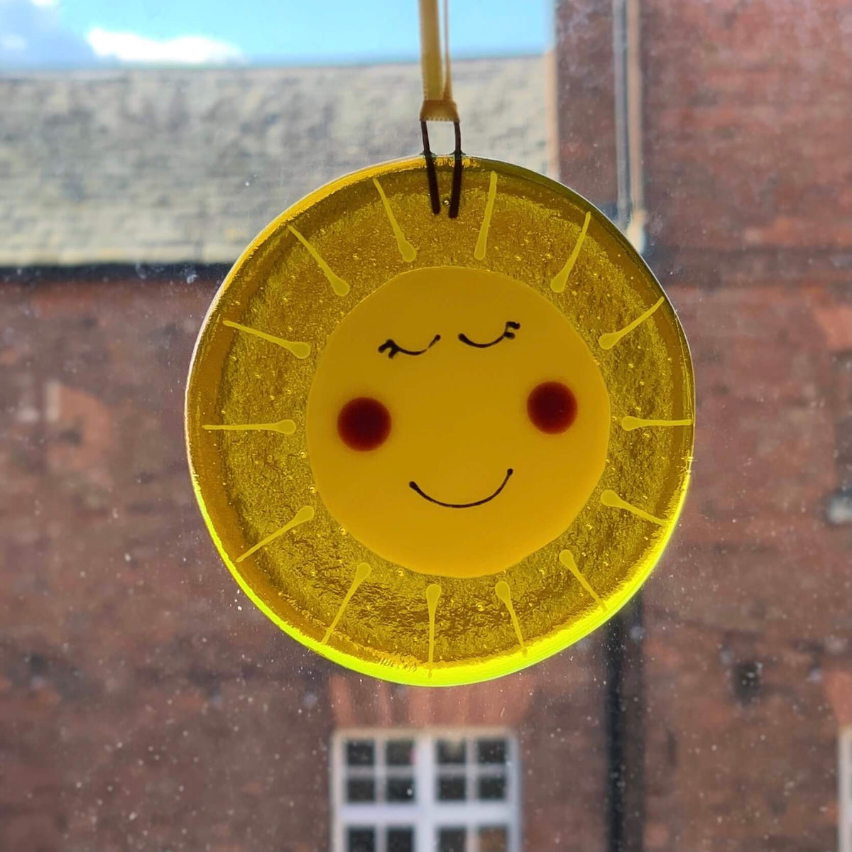 Handmade fused glass yellow circle hanging, with a smiley faced sunshine on the front, with rosey cheeks. Hangs from a piece of yellow ribbon in the window.