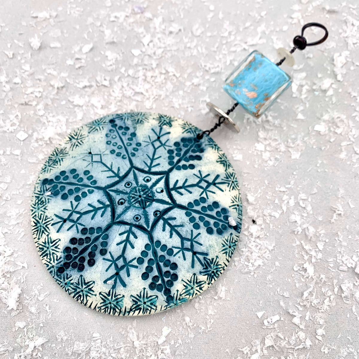 Handmade ceramic snowflake hanging decoration by Shirley Vauvelle. Featuring a handmade ceramic disk, with a snowflake pattern printed on with blue glazing, hanging from a thick wire with a blue button. Made in the uk.